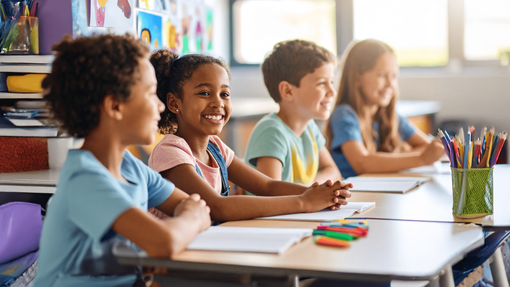 Vier Grundschulkinder sitzen gemeinsam an einem Tisch im Klassenzimmer, lächeln und wirken aufmerksam und fröhlich. Im Hintergrund sind bunte Kunstwerke und Schulmaterialien zu sehen – eine lebendige, positive Lernumgebung.