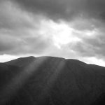 Sonnenstrahlen fallen durch Wolken auf dunkles Bergmassiv und schaffen Ruhe.