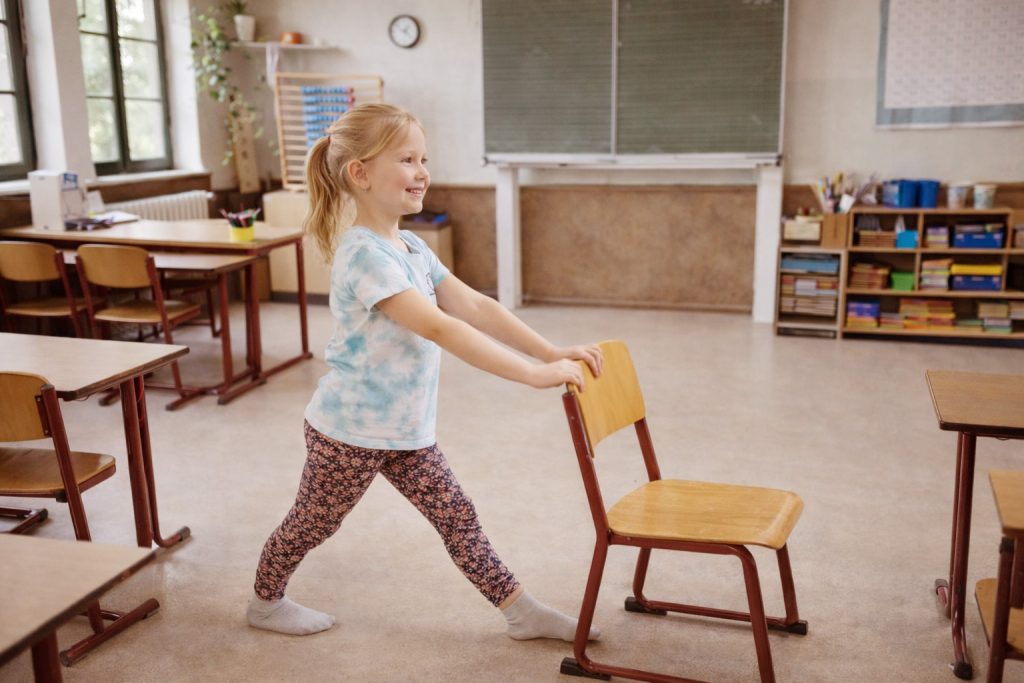 Kind macht spielerische Yoga Bewegung mit Stuhl im Klassenraum waehrend des Schulalltags